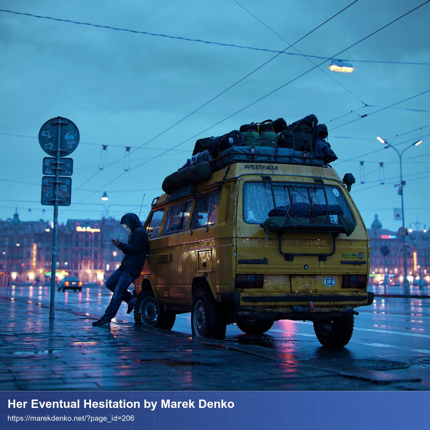 Eine Person lehnt gegen einen gelben VW Bus auf dessen Dach viel Gepäck verstaut ist. Das Wetter ist regnerisch blau. Im Hintergrund warme Lichter der Fenster der Stadt.
