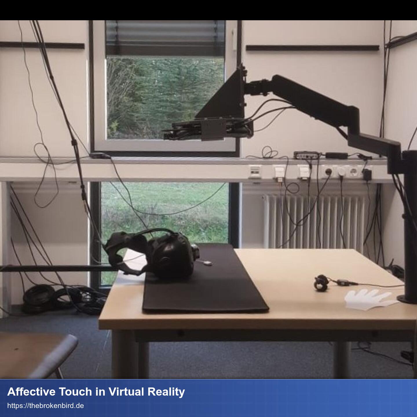 Ein Tisch von der Seite. Auf dem Tisch liegt ein VR-Headset und am Tisch ist ein Aufbau mit einem mechanischen Arm, an dessen Ende das UltraSound-Board befestigt ist und zur Tischplatte zeigt.