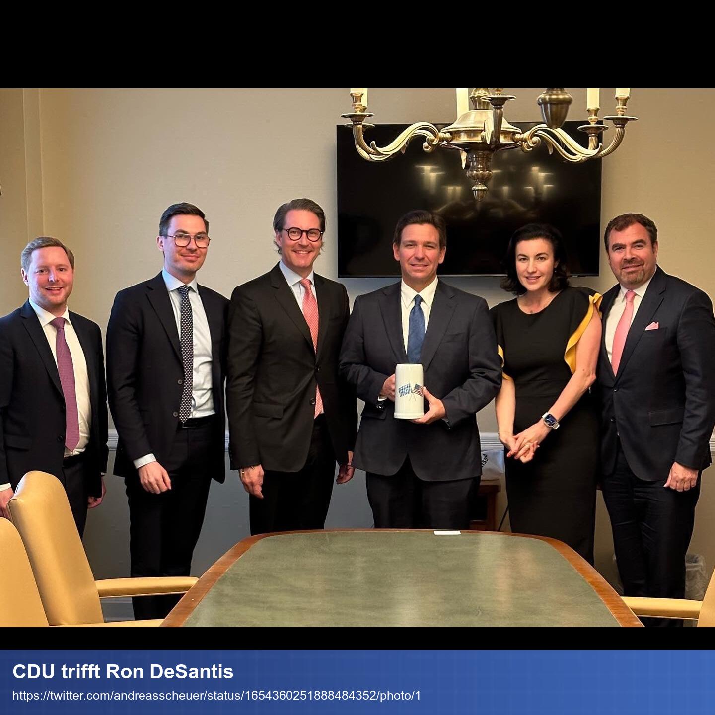 Scheuer und andere CDU Politiker stehen mit Ron DeSantis hinter einem Tisch, schauen in die lächelnd in die Kamera und Desantis hält einen kleinen Bierkrug in die Kamera.